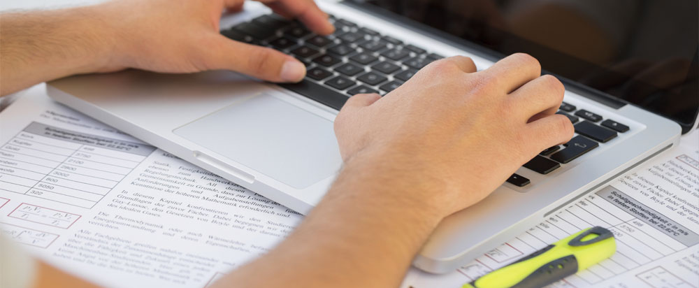 student typing on a computer with documents and a highlighter on the table in front of him